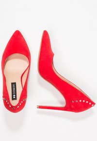 Talons hauts en daim rouge avec des bouts pointus, présentant des accents cloutés près du bout et un talon fin. Texture lisse et design élégant.