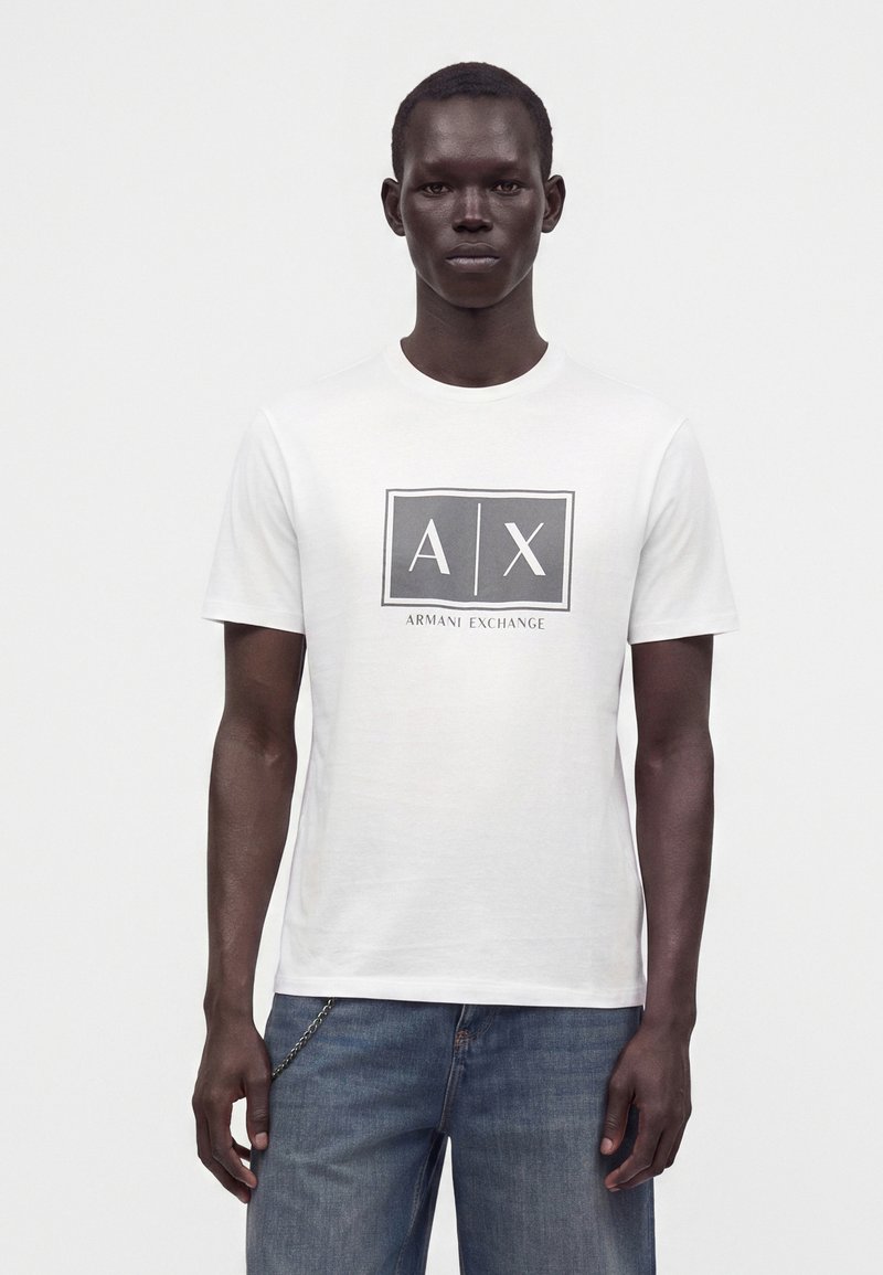 Άνδρας που φοράει λευκό T-shirt Armani Exchange με το λογότυπο AX και μπλε τζιν, στέκεται μπροστά σε απλό λευκό φόντο.