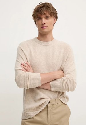 Beige knitted sweater with a round neckline and ribbed cuffs, featuring a relaxed fit and side slits, paired with light tan trousers.