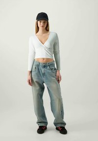 Blusa branca de manga longa em estilo wrap e jeans largos desbotados de tom azul claro, combinados com um boné preto e ténis pretos com detalhes vermelhos.