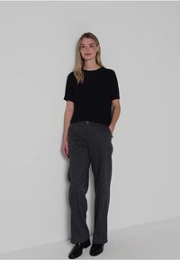 Černý top s krátkým rukávem v kombinaci se šedými volnými kalhotami. Hladký materiál; černé boty. Minimalistický design na světle šedém pozadí.