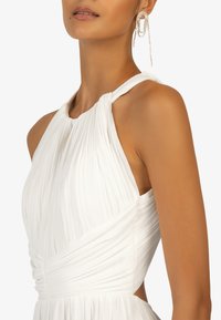 Biała sukienka z plisowanym materiałem, z dekoltem typu halter i dopasowaną talią. Posiada gładką, miękką teksturę i elegancki detal w postaci drapowania. Uzupełniona zwisającymi kolczykami.