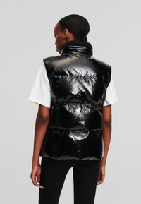 Vest puffer negru cu o textură lucioasă, guler înalt și design fără mâneci. Are secțiuni matlasate și un croi lejer.