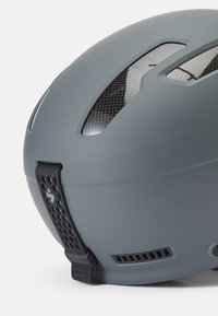 Casco de ciclismo gris con acabado mate, forma aerodinámica y ranuras de ventilación de fibra de carbono negra. Presenta una correa para el mentón texturizada y un logotipo.