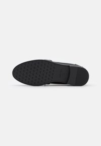 Black shoe bottom with textured grip pattern, rubber sole, flag logo detail, and slight curvature for stability. Smooth edges and no visible wear.