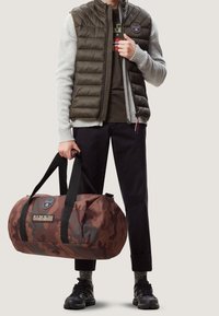 Gefütterte olivgrüne Weste über einem grauen Pullover, schwarze Hose und schwarze Sneaker. Hält eine braune Camouflage-Duffel-Tasche mit Logoplatte.