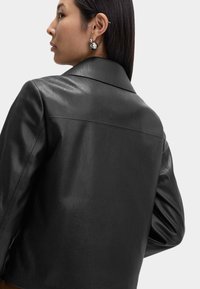 Kvinde med glat, mørkt hår, iført en sort læderjakke og sølv kugleøreringe, vist fra ryggen og siden mod en hvid baggrund.