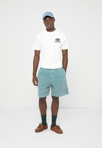Young man wearing a white graphic T-shirt, teal shorts, blue cap, green socks, and brown boat shoes standing against a white background.