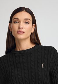 Svart kabelstickad tröja med ribbad halsringning; har en liten broderad logo i kontrastfärg på bröstet.