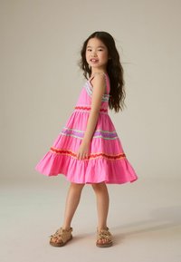 Jeune fille aux longs cheveux foncés portant une robe rose sans manches à rayures colorées, debout et souriante sur un fond uni.