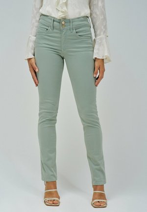 Jeans skinny verde claro con una textura suave, cintura alta, dos bolsillos frontales y detalles de botones plateados, combinados con una blusa blanca con volantes.