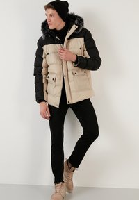 Giacca a vento beige e nera con cappuccio foderato in pelliccia, dotata di due tasche frontali e chiusura con zip, indossata con pantaloni neri e sneakers beige.