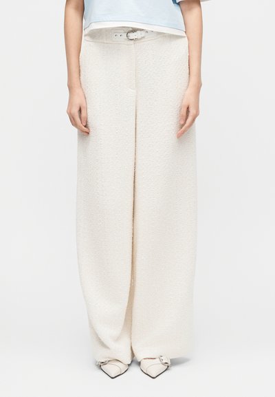 Λευκό υφασμάτινο παντελόνι φαρδιάς γραμμής με θηλιές για ζώνη, συνδυασμένο με λευκά μυτερά παπούτσια με λεπτομέρεια αγκράφας, φορεμένα από ένα όρθιο άτομο.