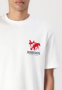 Hvid bomulds T-shirt med en rød broderet grafik af en cowboy på en hest og blå tekst, der læser "RODEO DAYS." Enkel, afslappet design.