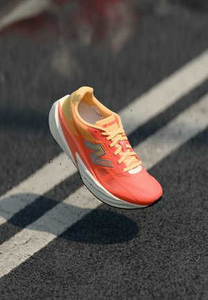 Chaussure de course orange vif et jaune avec le logo New Balance, suspendue au-dessus de l'asphalte avec des marquages de voie blancs.