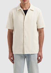 Kortarmig, lichtbeige overhemd met een gestructureerde wafelweving. Voorzien van een knoopsluiting en een klassieke kraag. Gecombineerd met lichte denim jeans.