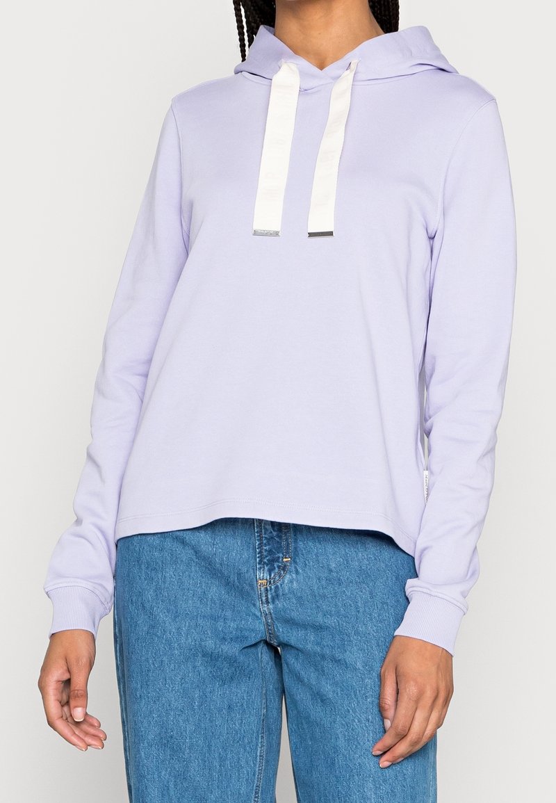 Femme portant un sweat à capuche violet clair avec des cordons blancs et un jean bleu, debout, les mains détendues le long du corps.