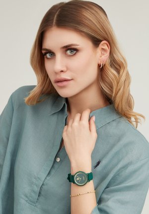 Donna con capelli biondi che indossa una camicia verde acqua con colletto, orecchini a cerchio dorati, orologio da polso verde e delicato braccialetto d'oro al polso sinistro.