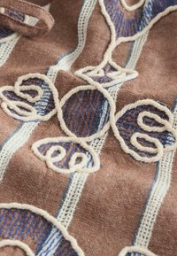 Tissu marron avec des rayures verticales crème et bleues, présentant une broderie en fil crème en relief délimitant des formes de feuilles abstraites.