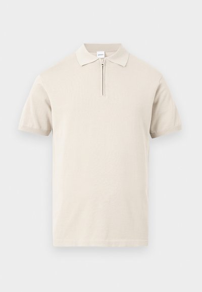 Polo beige à manches courtes avec col rabattu et courte fermeture éclair frontale sur un fond uni.