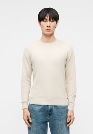 Giovane uomo con capelli neri e lisci che indossa un maglione beige a maniche lunghe e jeans blu, in piedi di fronte a uno sfondo bianco.