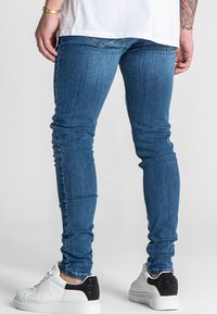 Jeans de mezclilla azul con un ajuste slim, que presentan un lavado desgastado y un diseño de cinco bolsillos, combinados con zapatillas blancas con detalles negros.