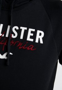 Svart hoodie med "Hollister California" broderat i vitt och rött. Inkluderar snodd och en mjuk textur.