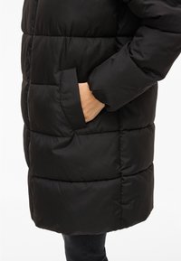 Crna jakna s prešanim dizajnom, s bočnim džepom i glatkom teksturom. Jakna ima opušteni kroj i do koljena dosezajući stil.