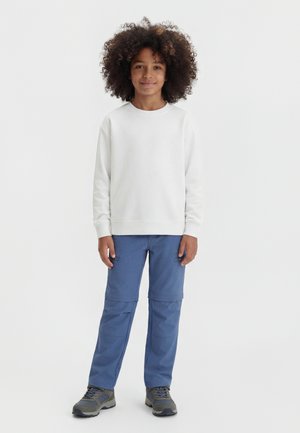 Niño con cabello rizado que lleva una camiseta blanca de manga larga, pantalones convertibles azules y zapatillas grises, de pie contra un fondo blanco liso.