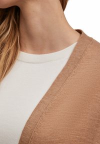 Beige Cardigan aus weichem Stoff, mit V-Ausschnitt und einem cremefarbenen Top darunter. Der Cardigan weist subtile Texturdetails auf.
