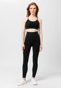 Zwarte cropped sportbeha met dunne bandjes, gecombineerd met hoge zwarte leggings. Model draagt witte sneakers en staat op een neutrale achtergrond.