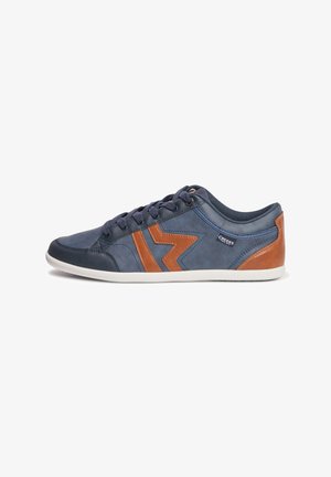 Zapatilla de color azul marino con acentos de cuero marrón, que presenta un diseño de zigzag prominente, parte delantera con cordones y suela de goma blanca. Textura suave.
