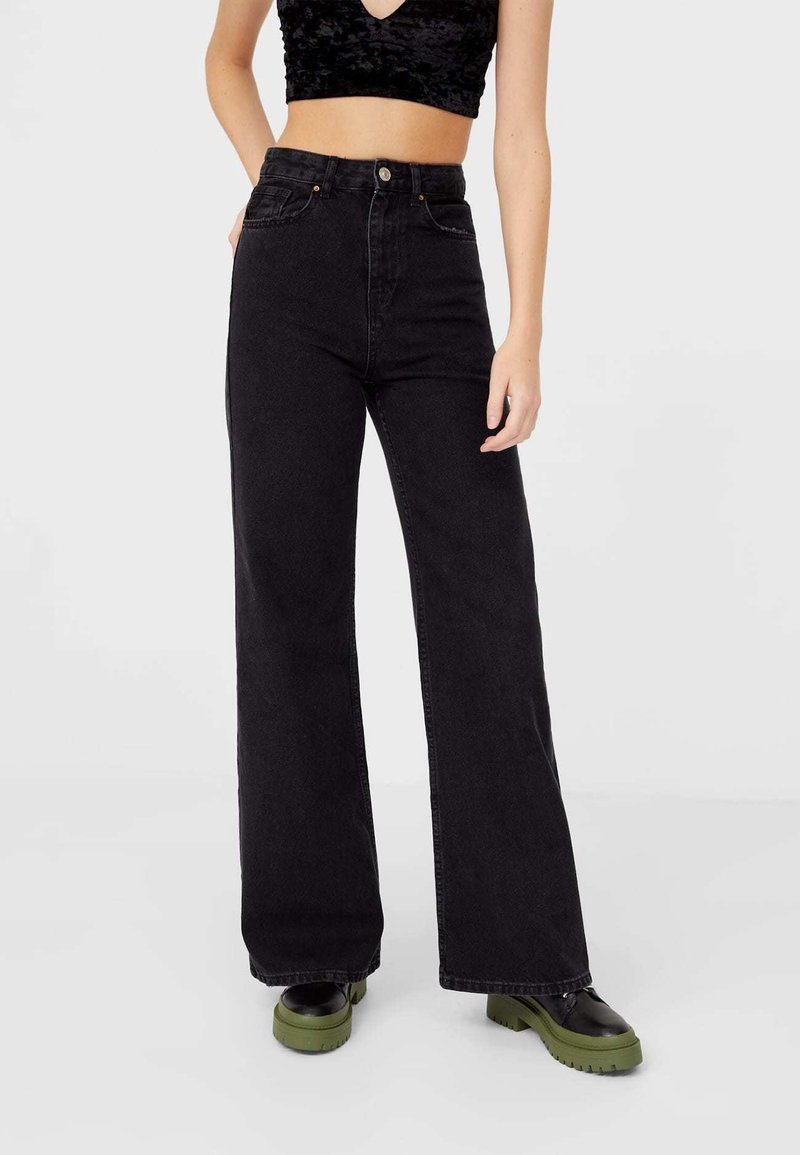 Femme portant un jean noir taille haute à jambes larges, assorti d'un crop top noir et de chaussures noires aux semelles épaisses vertes, debout devant un fond blanc.