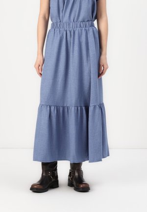 Frau mit blauem stufigem Midirock mit gerafftem Bund und schwarzen Stiefeletten mit Schnallen, stehend vor einem schlichten weißen Hintergrund.