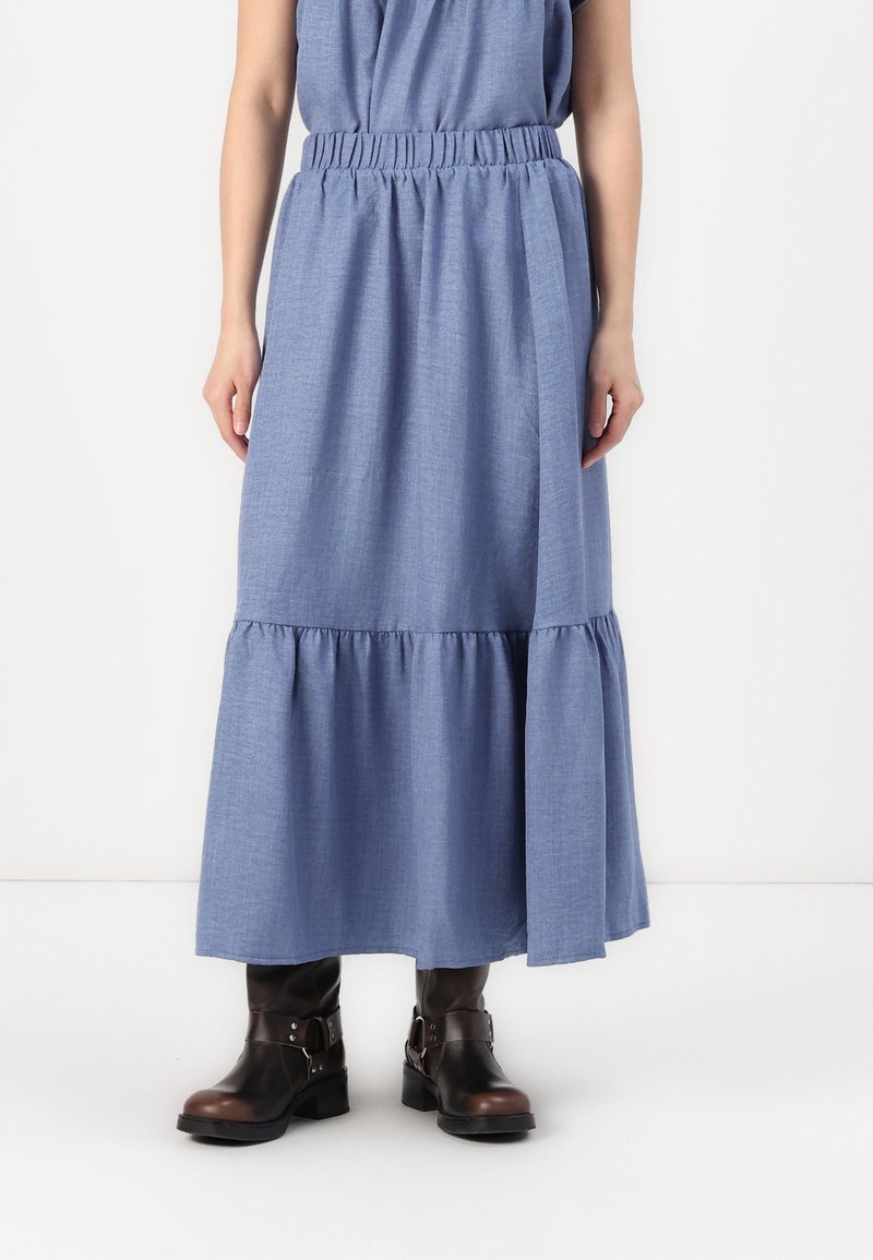 Femme portant une jupe midi bleue à volants avec une taille froncée et des bottines noires à boucles, debout devant un fond blanc uni.