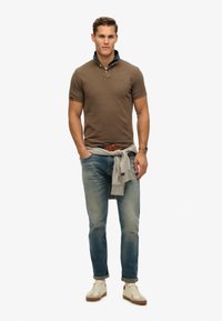 Polo marrone con colletto in denim, jeans azzurri chiari, maglione grigio annodato in vita e sneakers crema con suola in gomma.