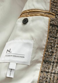 Šedá a hnědá texturovaná tkanina s krémovou podšívkou a hnědým akcentem, obsahující černý knoflík, štítek s nápisem "N. PREMIUM COLLECTION" a detaily šití.