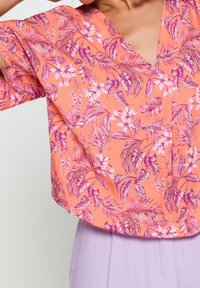 Blusa a manica corta in corallo con un motivo floreale caratterizzato da foglie e fiori rosa. Design a V, vestibilità drappeggiata, realizzata in tessuto leggero.