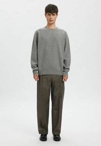 Hall et sügiskampsun ümmarguse kaelusega, pruunide ruuduliste pükste ja mustade kingadega, neutraalsel taustal.