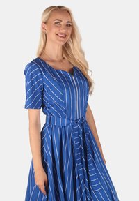 Blauriesiertes Kleid mit kurzen Ärmeln, einem V-Ausschnitt und einem taillierten Oberteil. Der Rock ist voluminös mit einem Taillengurt. Glattes Material und weiße Streifen.