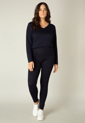 Vrouw met lang donker haar, staand, gekleed in een zwart shirt met lange mouwen, zwarte legging en witte sneakers tegen een effen lichte achtergrond.