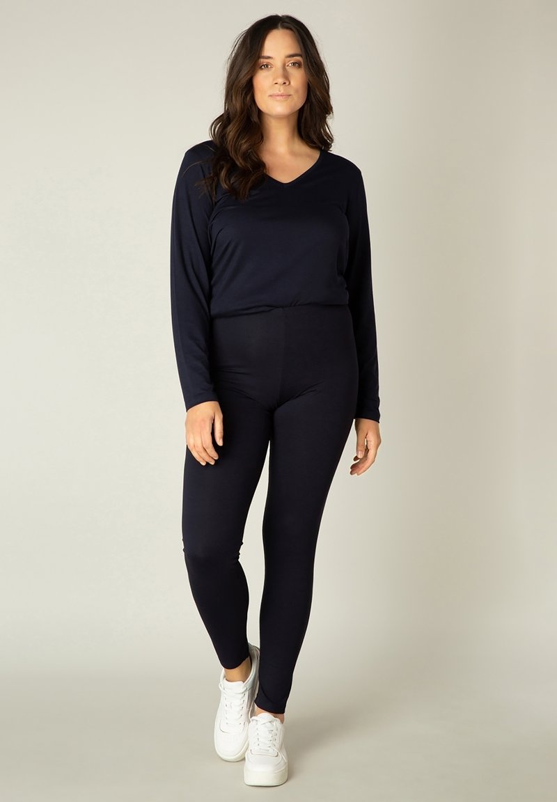 Vrouw met lang donker haar, staand, gekleed in een zwart shirt met lange mouwen, zwarte legging en witte sneakers tegen een effen lichte achtergrond.