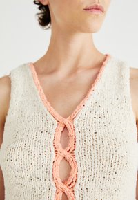 Gebreide tanktop met een crèmekleurige textuur en een V-halslijn, accentueert met een gevlochten perzikkleurige rand en een sleufdetail in het midden.
