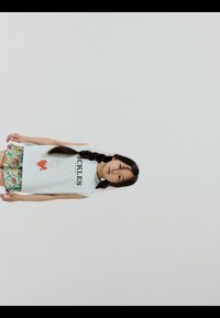 Niña con cabello largo trenzado, vestida con una camiseta blanca sin mangas con un diseño floral y pantalones cortos de colores con estampados, sobre un fondo liso.