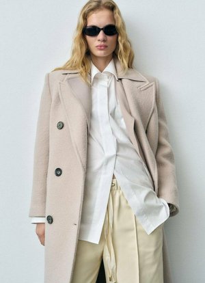Beige wool coat with a structured design, black buttons, layered over a white shirt. Notable for its relaxed fit and sleek beige trousers.