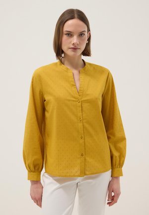 Mujer con blusa de botones color mostaza con textura sutil de puntos y pantalones blancos, de pie contra un fondo claro y sencillo.