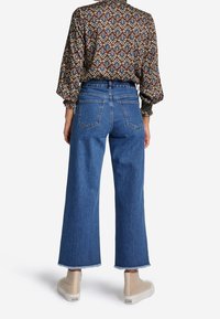 Hoch taillierte, weit geschnittene blaue Jeans aus Denim mit fransigem Saum, kombiniert mit einem gemusterten Langarmoberteil und beigen High-Top-Sneakern.