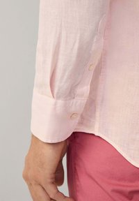 Svetloružová ľanová košeľa s jemnou textúrou, s gombíkovým manžetou a mierne prehnutým spodným okrajom. Ruka položená na ružovom textile.
