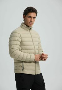 Beige pufferjack met een hoge kraag, ritsluiting en een horizontaal gestikt patroon. Heeft zijzakken en een zachte, glanzende textuur.