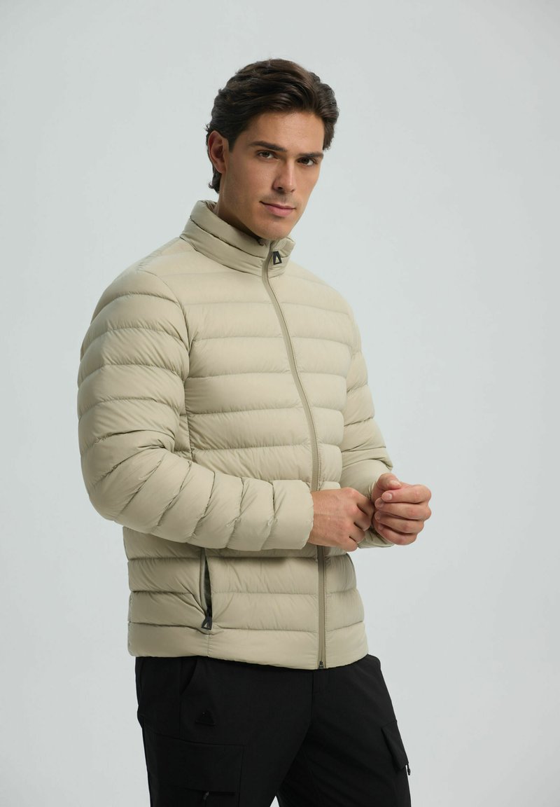 Beige pufferjack met een hoge kraag, ritsluiting en een horizontaal gestikt patroon. Heeft zijzakken en een zachte, glanzende textuur.